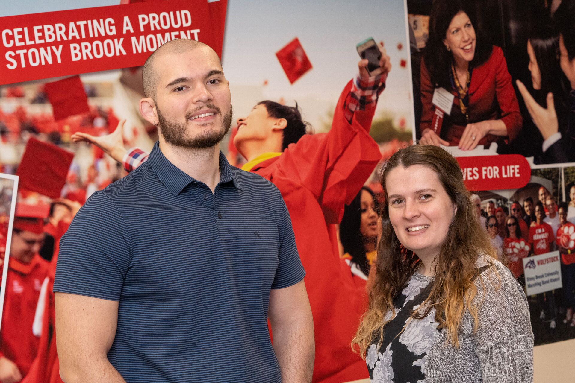 Young white male with mostly shaved head and short beard wearing a striped short sleeve golf shirt (Vincent) on left next to a young white woman with long light brown hair, shirt and sweater (Sherie) both smiling in front of an SBU wall banner