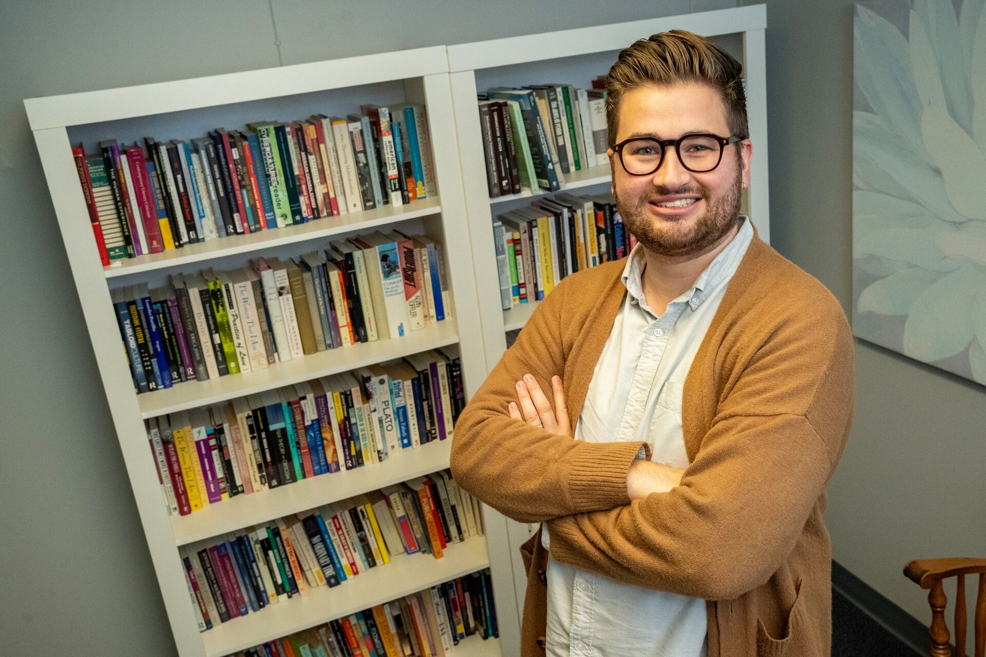 Caucasian man with facial hair and glasses, sweater and dress shirt, standing arms folded in front of a bookshelf