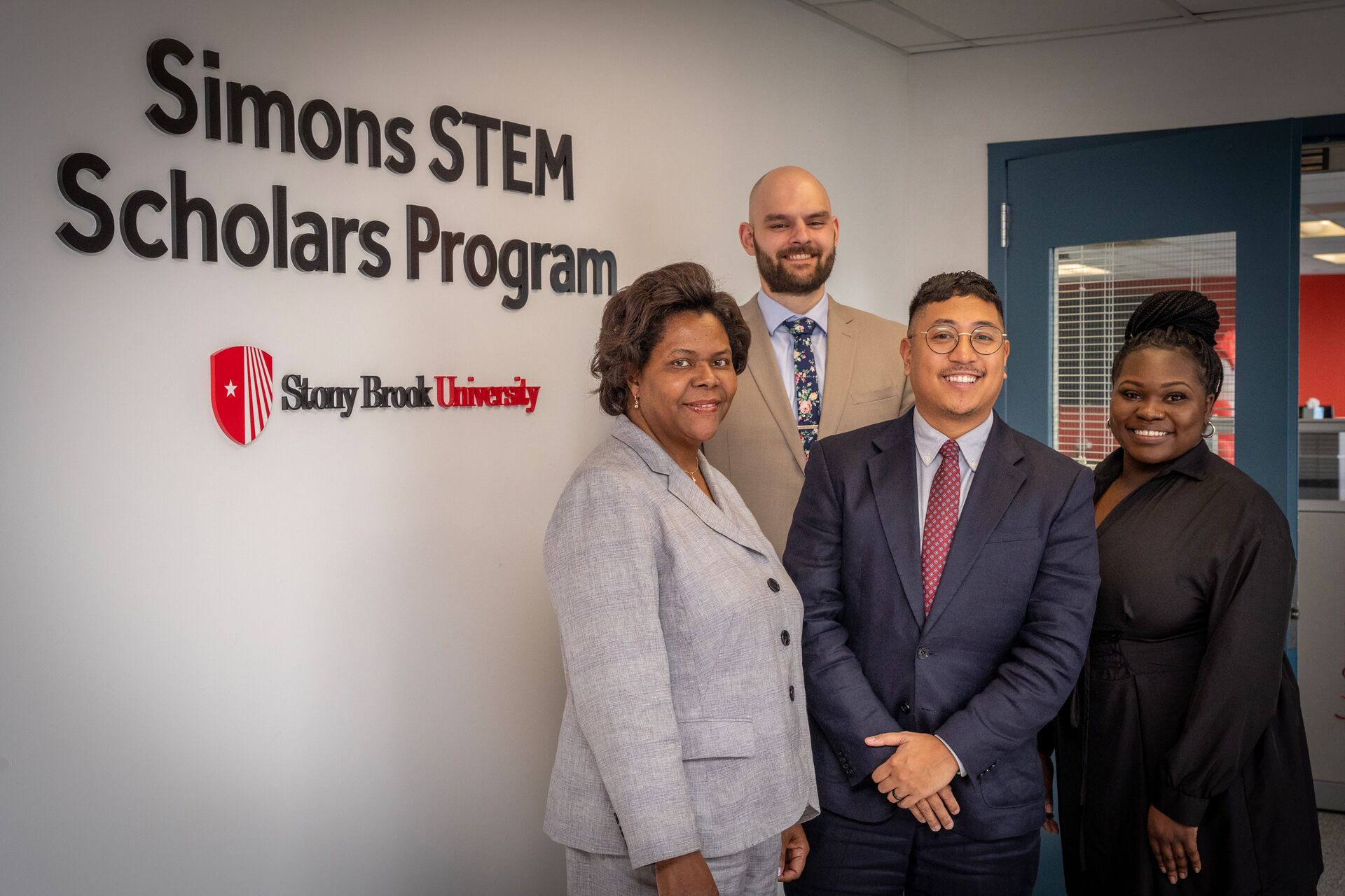 Two women and two men in professional attire standing together and smiling in front of a white wall that displays the "Simons STEM Scholars Program" and Stony Brook University logo.