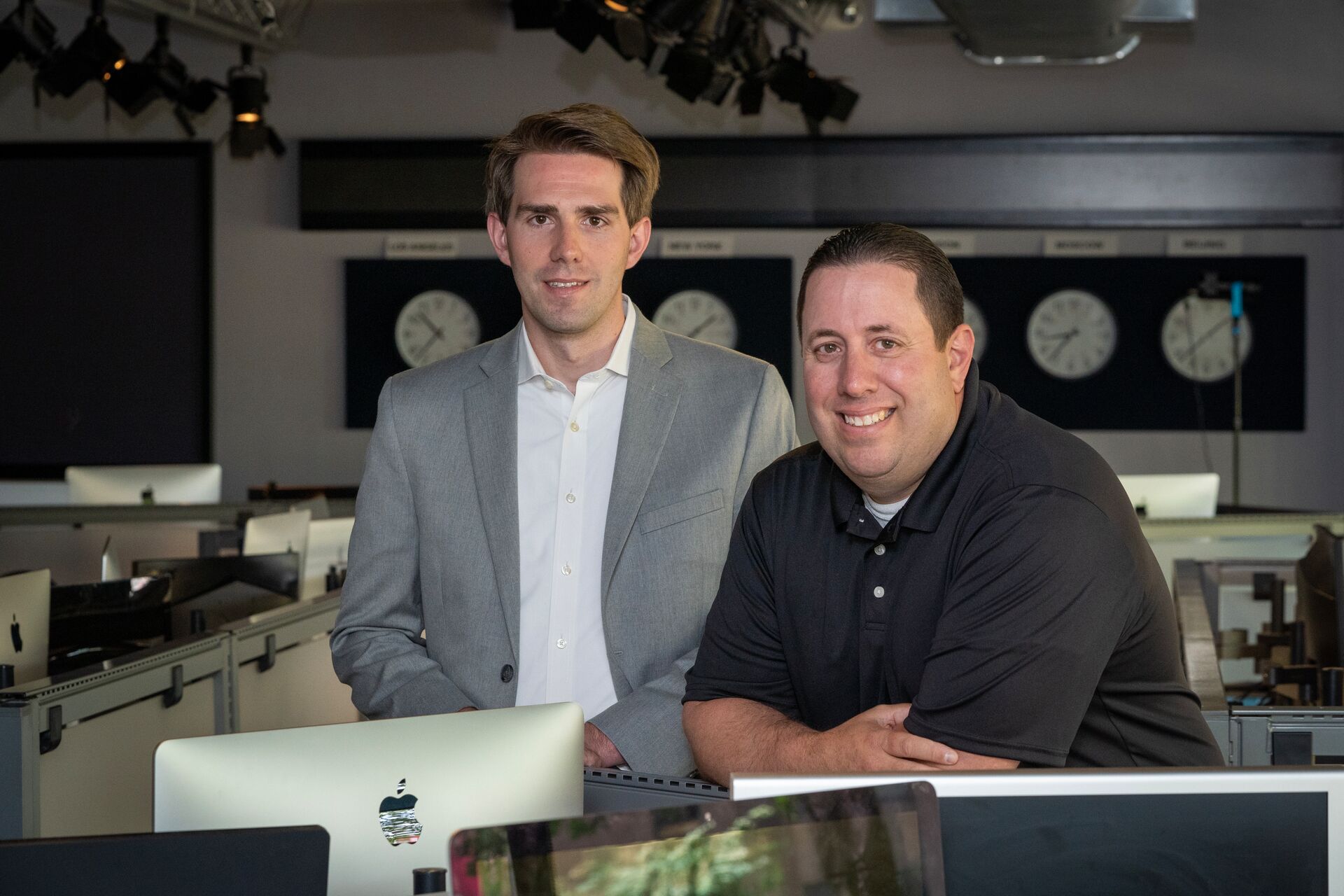 Two white men -- one with light brown hair (Evan Tousey, on left) wearing a suit jacket and shirt, the other with very short black hair and a shortsleeved collared shirt (Eric Brodsky), smiling and leaning on a computer table