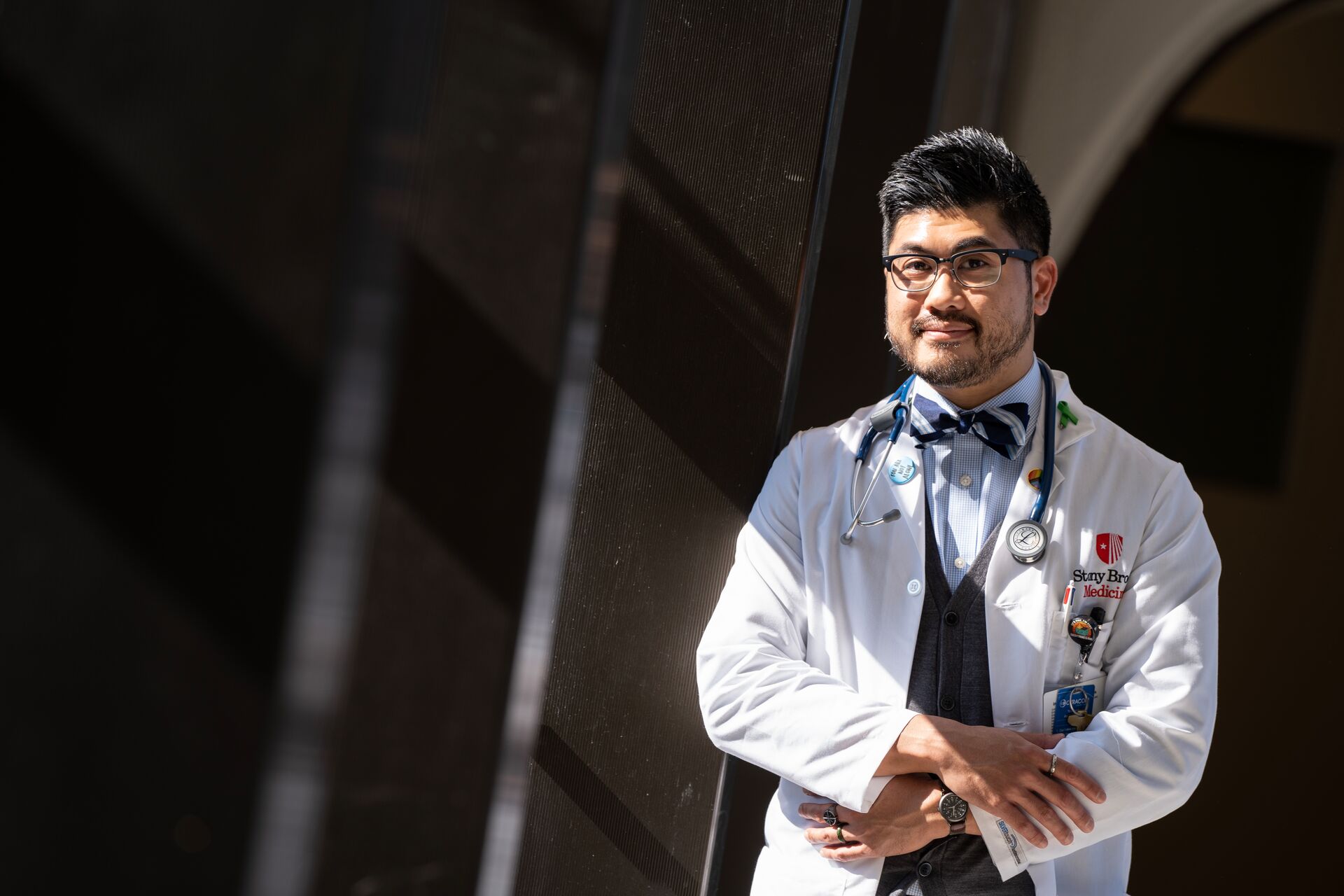 Asian man with facial hair and glasses, wearing a white medical coat with shirt and bowtie underneath and a stethoscope around his neck (Andrew) standing and smiling