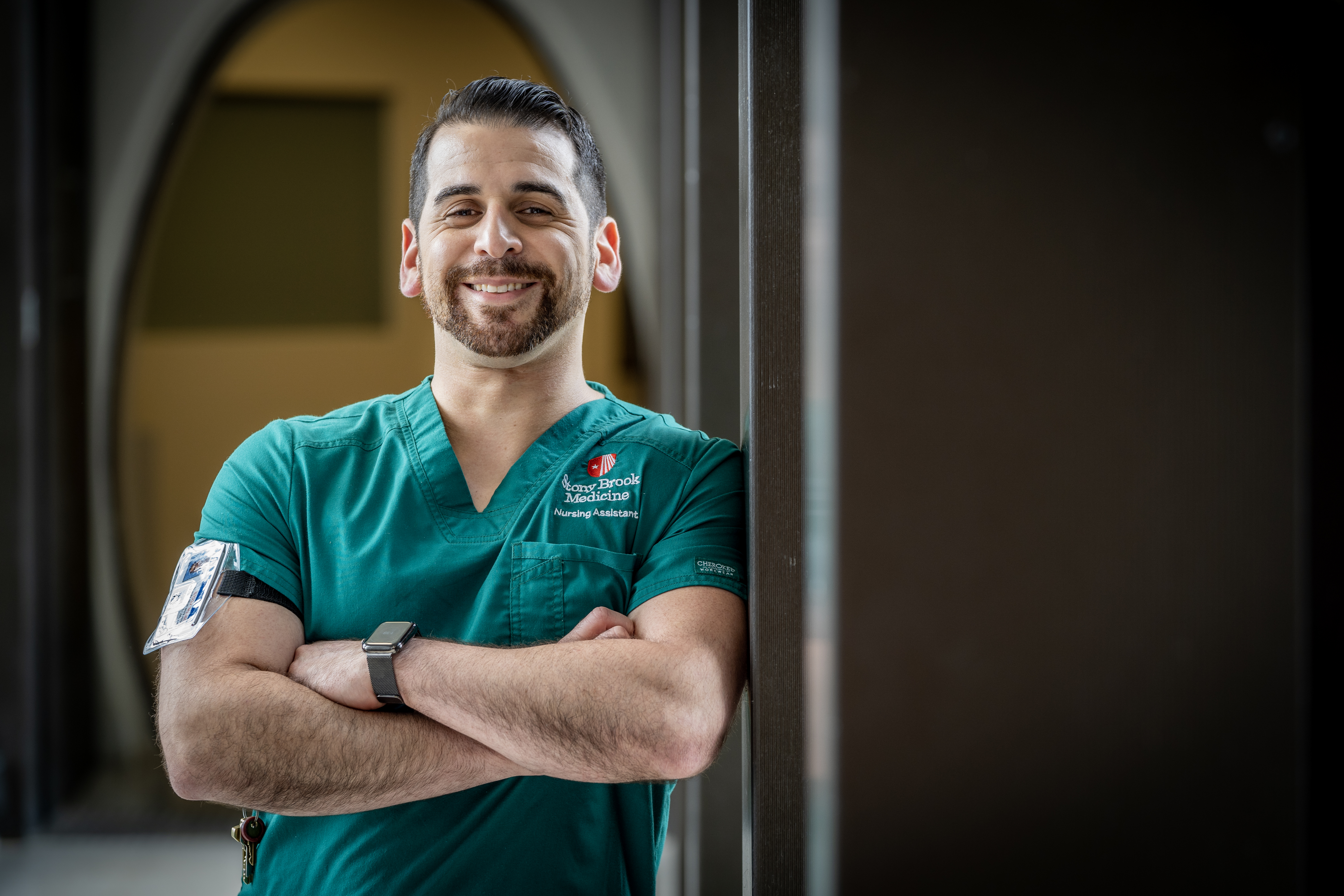Caucasian man with facial hair wearing green scrubs, arms crossed across chest and wearing a watch