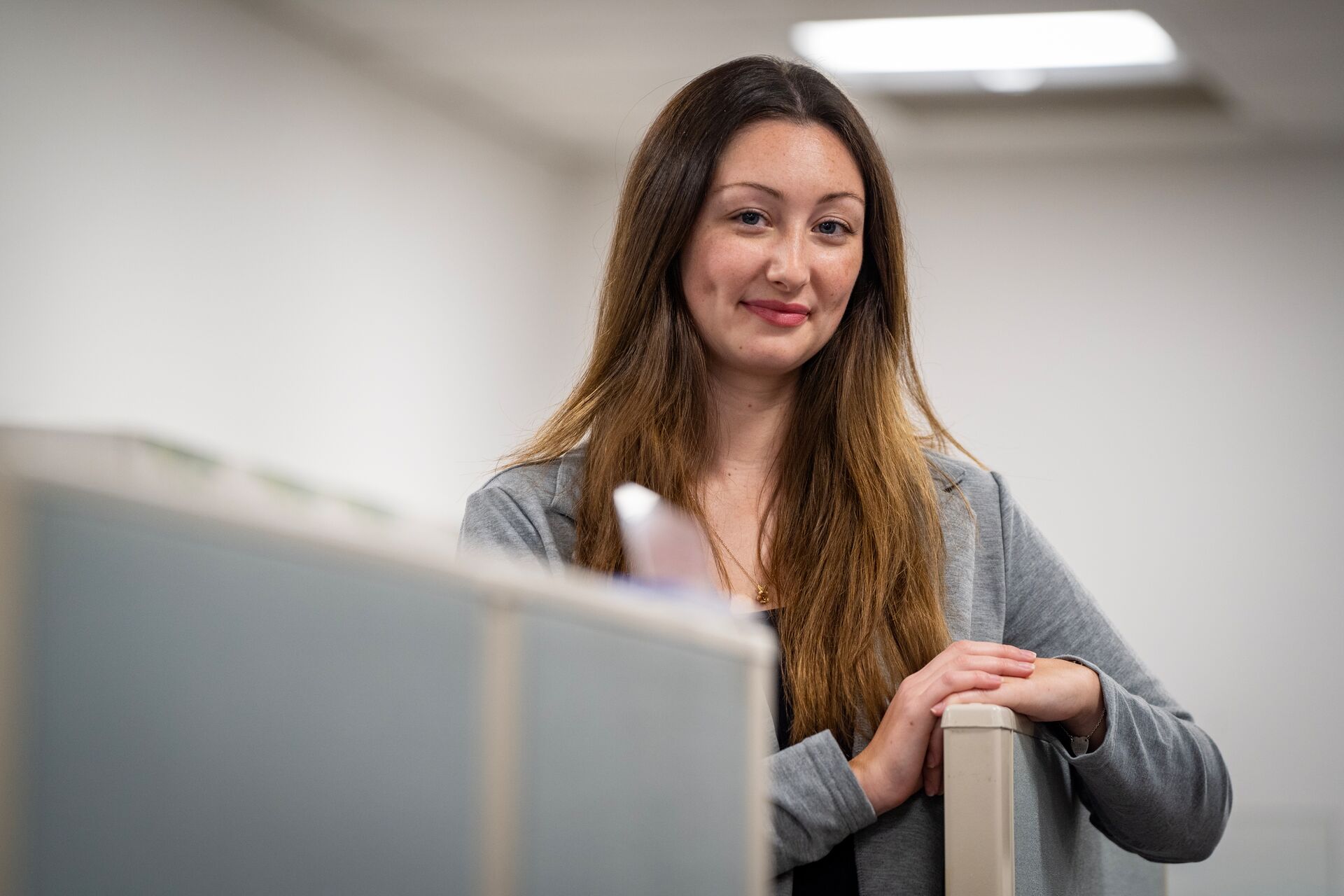 Caucasian woman with long brown hair, wearing gray jacket and standing behind cubicle