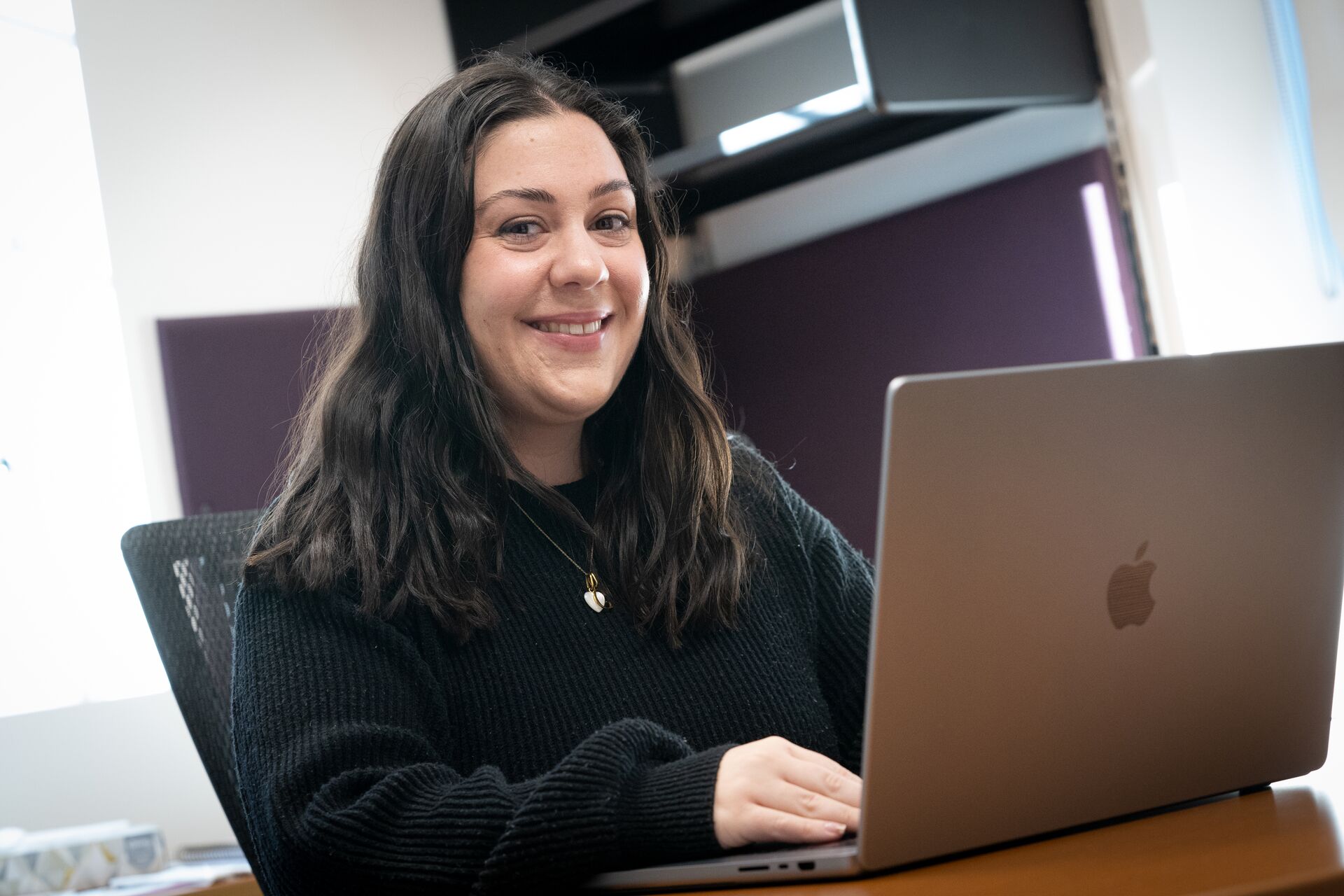 Caucasian woman with long black hair wearing black long-sleeved shirt, sitting in front of open laptop)