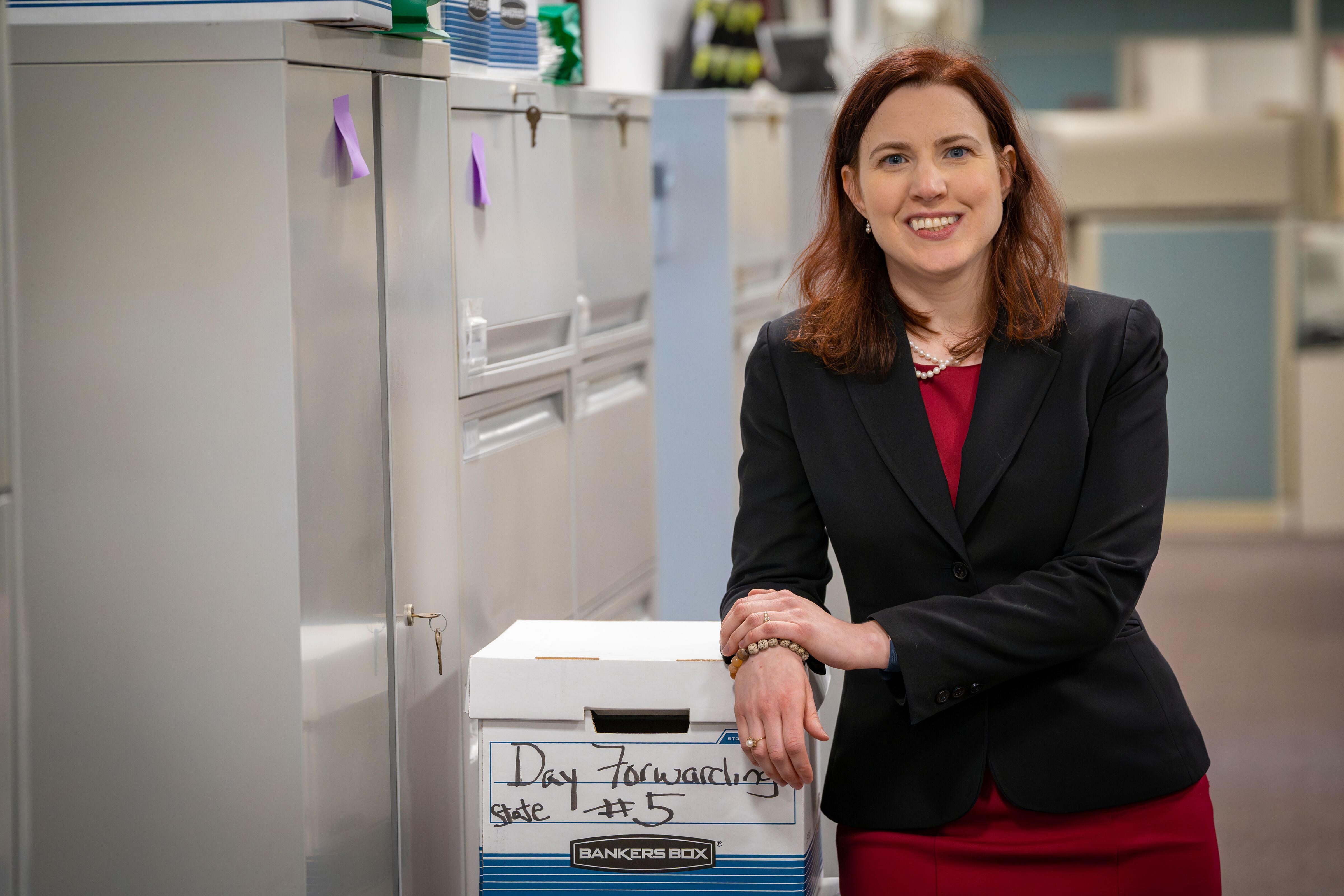 Caucasian woman with midlength red hair wearing black blazer (buttoned) over red dress and leaning against a file cabinet