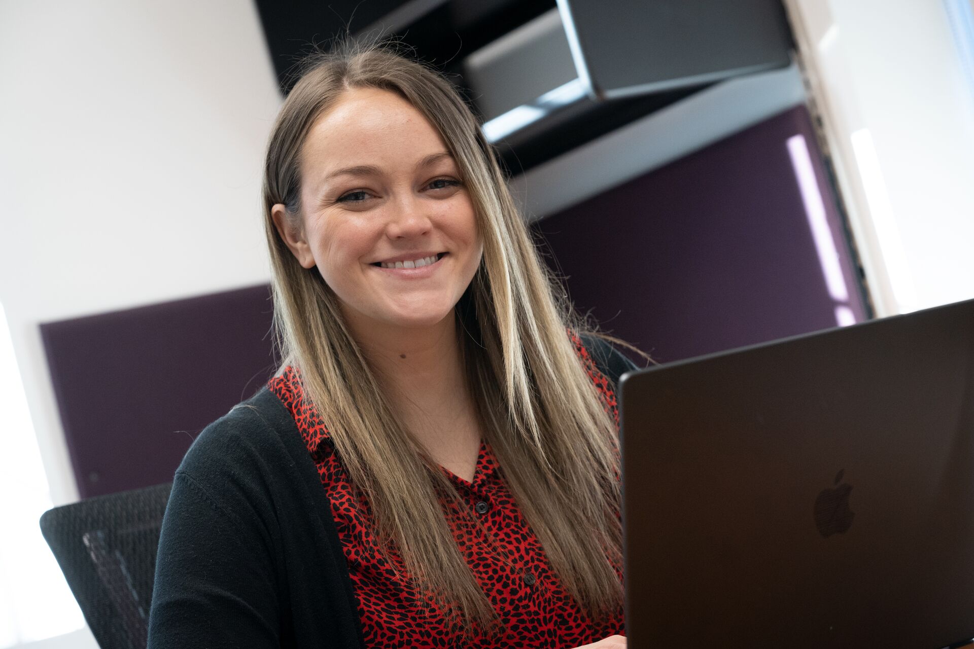Caucasian woman with long blonde hair wearing red shirt with black cardigan, sitting in front of open laptop
