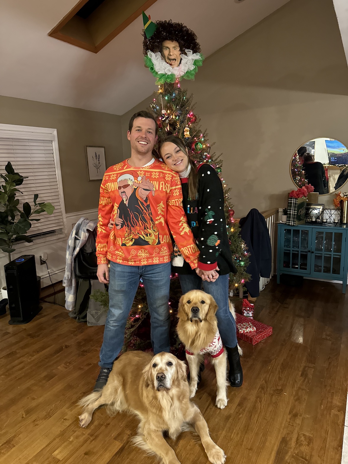 Caucasian couple (male and female) standing in front of Christmas tree with two golden retrievers on the ground beside them