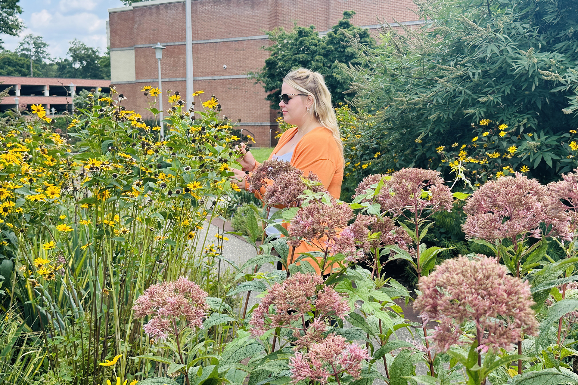 Blonde caucasian woman wearing sunglasses and orange blouse standing in some landscaping