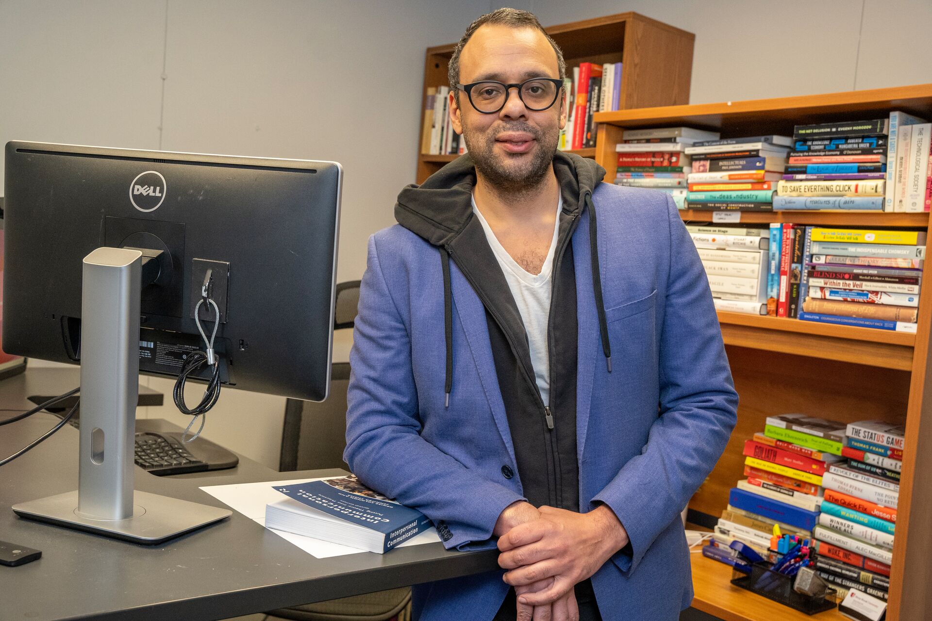 Caucasian man with facial hair and glasses wearing blue hoodie standing in front of bookshelf and leaning on computer desk 