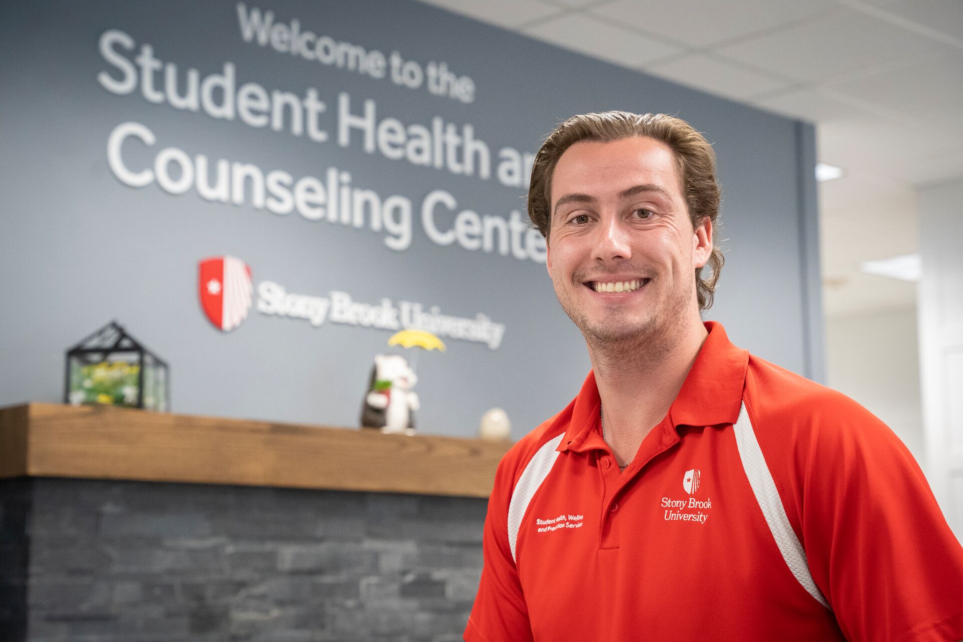 Blonde caucasian man wearing red athletic t-shirt, in front of desk with sign behind it that says, "Welcome to the Student Health and Counseling Center"