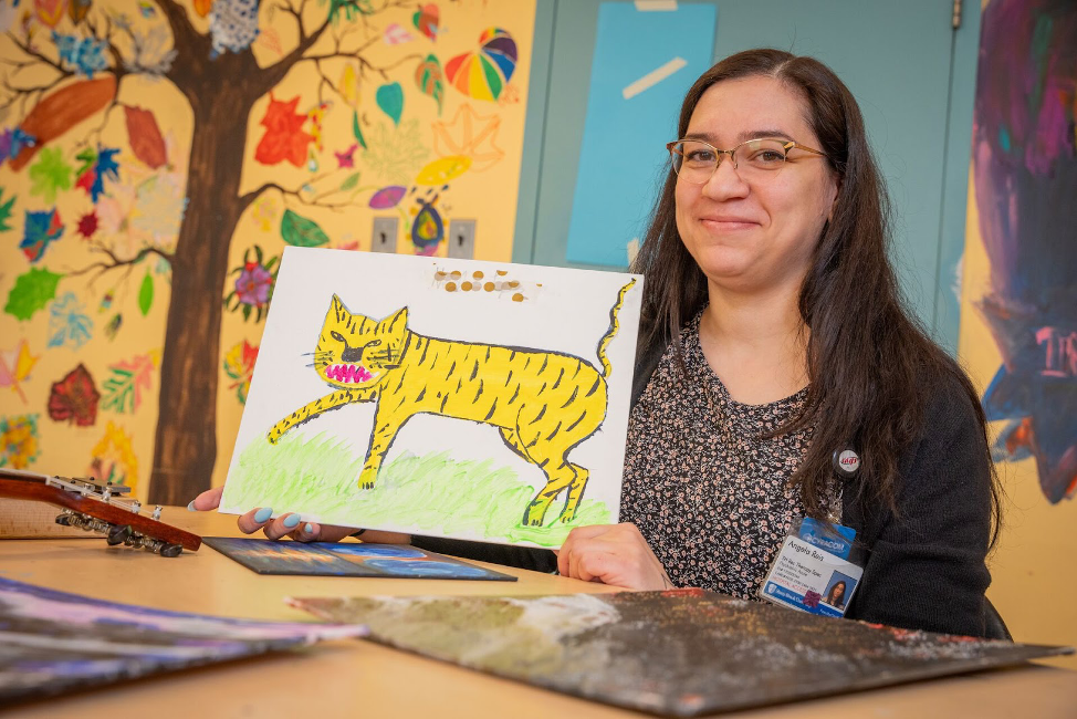 Young woman with long black hair and glasses smiling and holding up a drawing of a tiger, with a colorful tree and leaf drawn mural behind her
