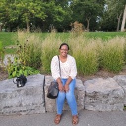 African American woman wearing white long sleeved blouse, jeans and black bag; sitting on large garden stones with landscaping behind her.