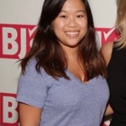 Asian American woman hair a couple inches past her shoulders, wearing gray short sleeved t-shirt, standing in front of a step-and-repeat that has the BJ's logo and someone is standing to her right, but that person is cropped out