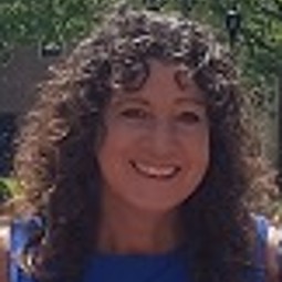 Middle aged caucasian woman with tight curls and bangs, wearing blue sleeveless blouse, photo cropped tight to face