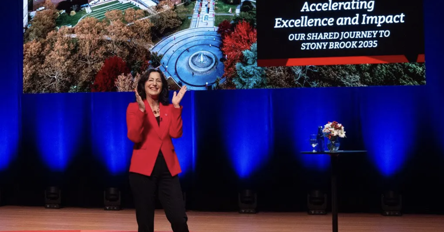 Woman with brown hair wearing red blazer and black pants on stage clapping