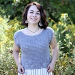 Caucasian woman with dark brown shoulder length wavy hair wearing purple knit short-sleeve shirt and purple and white skirt, standing in front of foliage
