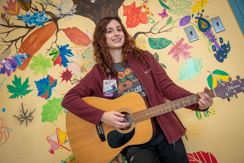 Young woman with long light brown hair smiling and playing the guitar with a colorful painted tree and leaf mural behind her