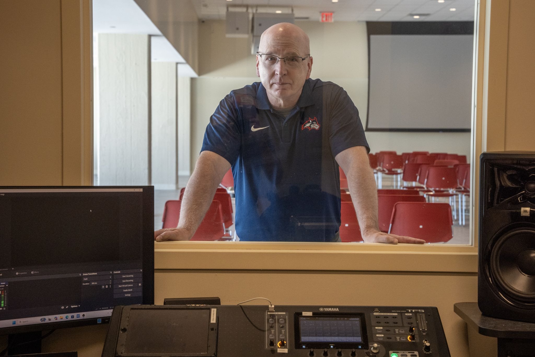 A man (Joe Kuhl), with glasses and a blue polo shirt looking through a window into an audio control room