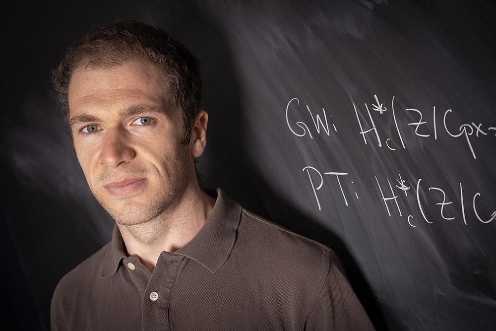 A man (John Pardon), in a brown polo shirt stands in front of a blackboard covered in mathematical equations.