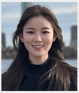 Close-up portrait of a smiling person with long dark hair wearing a black top, photographed outdoors against a blurred city background.