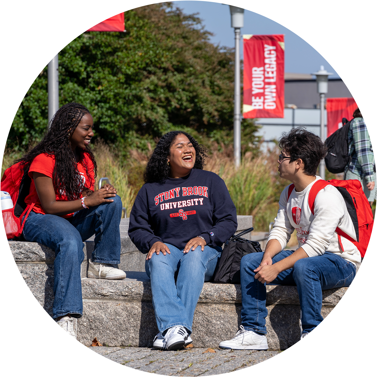 Three students sitting and talking outside on a stone ledge, with red banners in the background