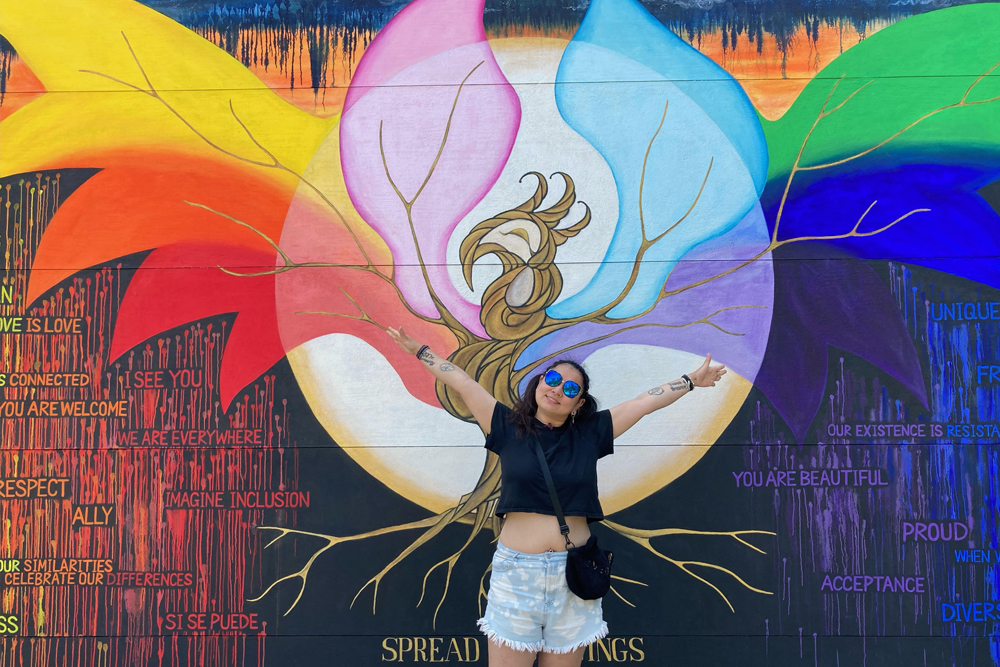 student standing infront of mural with hands spread, places in a way that the spread wings look it's coming from her