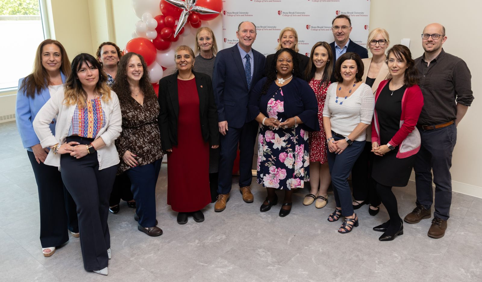 A group of smiling people stand in front of a backdrop with the Stony Brook University College of Arts and Sciences logo flanked with red and white balloons;
