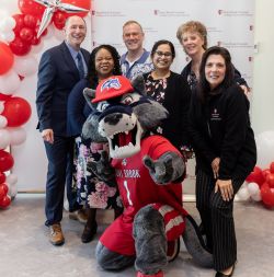 Six people stand in front of a backdrop with the Stony Brook University College of Arts and Sciences logo; balloons border the left side of the backdrop. Wolfie, the Stony Brook University mascot, kneels in front of the group. 