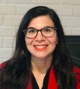 Portrait of a smiling person with glasses, wearing a blue top, set against a neutral background.