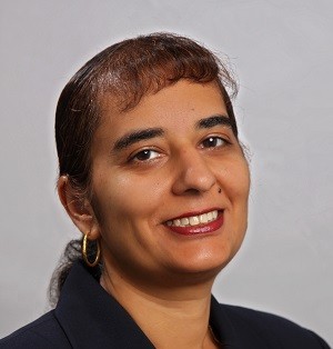 A woman in a black blazer smiles confidently against a gray background. She has medium-length hair, hoop earrings, and wears light makeup.