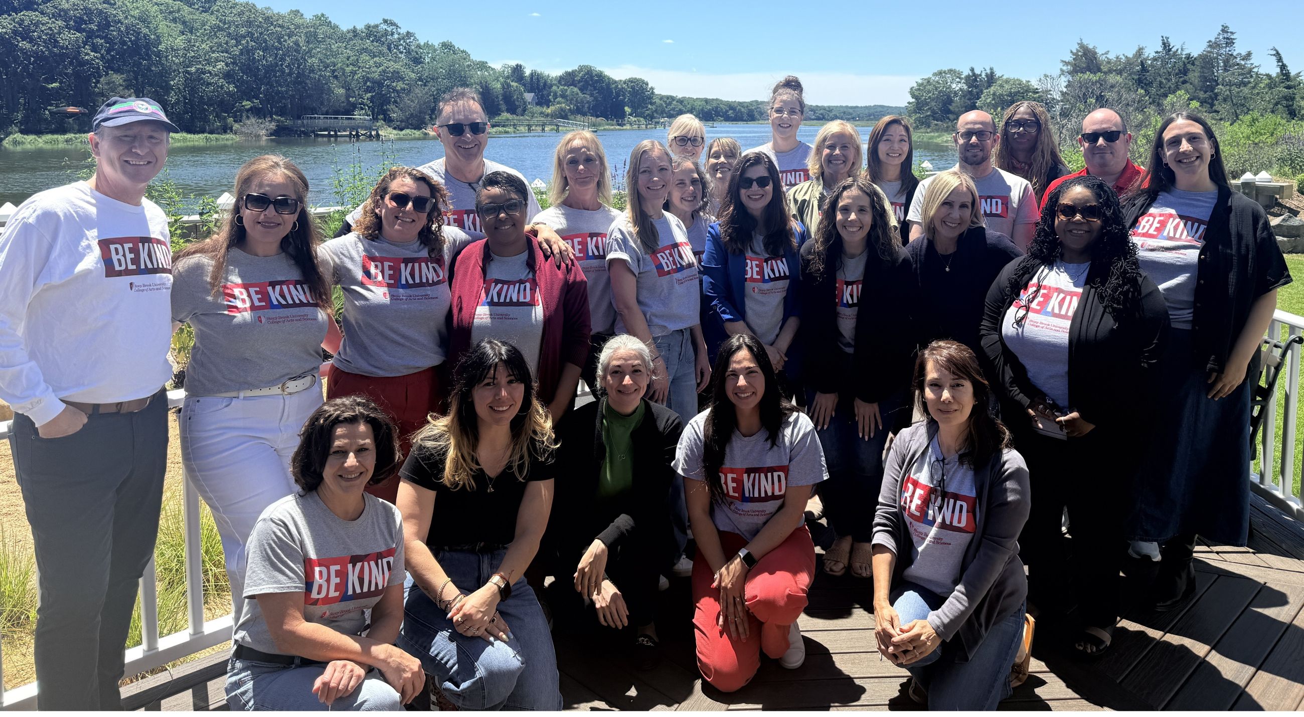 A large group of about two dozen smiling colleagues, many wearing matching gray "Be Kind" Stony Brook University College of Arts and Sciences t-shirts, pose together on a wooden deck by the water for a staff retreat photo. They stand and kneel in three rows on a sunny day, with a calm bay, a small dock, and tree-lined shoreline in the background.