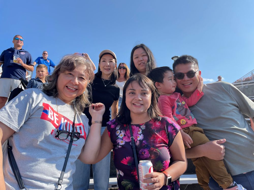 a group of smiling people stand in an outfoor stadium. One person wears a t-shirt that says Be Kind.