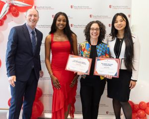 four people stand in front of a backdrop with the Stony Brook University College of Arts and Sciences logo; balloons are displayed at the top left. One person holds two certificates.