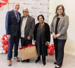 A group of four people stand in front of a backdrop with the Stony Brook University College of Arts and Sciences logo. One person holds a gift bag. 