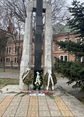 A memorial site with a tall stone monument flanked by white floral wreaths and ribbons. Flowers and photos are placed at the base, conveying respect and remembrance.