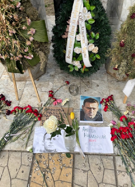 A memorial site with a tall stone monument flanked by white floral wreaths and ribbons. Flowers and photos are placed at the base, conveying respect and remembrance.