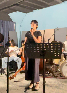 Person singing into a microphone with a band performing in the background in a room with overhead ductwork. Person singing into a microphone with a band performing in the background in a room with overhead ductwork.