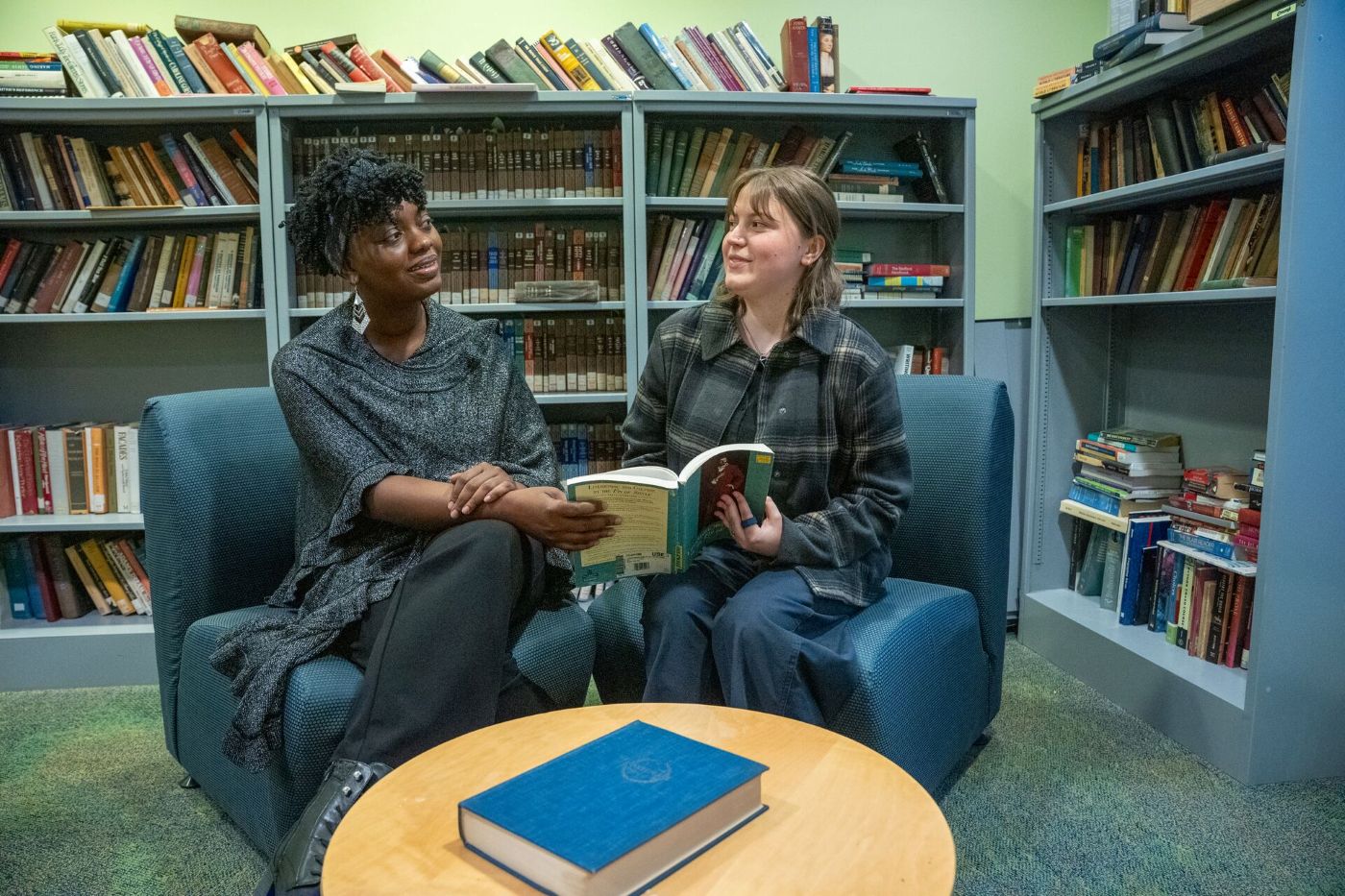 Two people sit in a library on blue chairs, holding books and conversing. Shelves filled with books line the background. A closed book is on the round table in front of them.