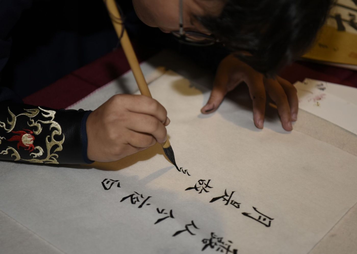 A person writing Chinese calligraphy with a brush on white paper, wearing a dark sleeve with embroidered patterns.