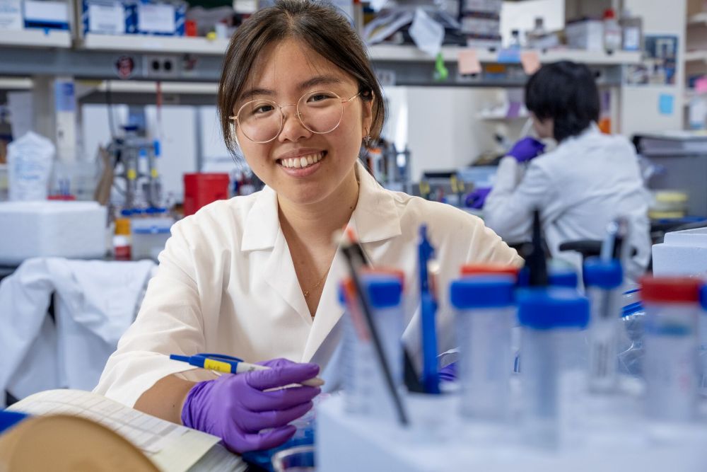 Person in a white lab coat and purple gloves smiling while holding a pipette at a laboratory workstation with equipment in the background.