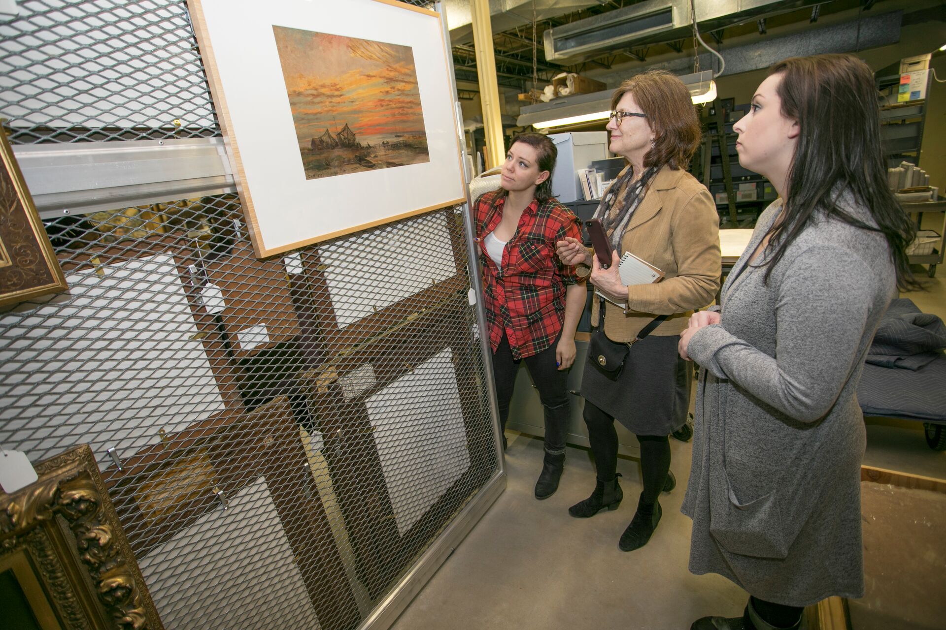 Three people examine a framed landscape painting stored on a mesh rack in an art conservation space.