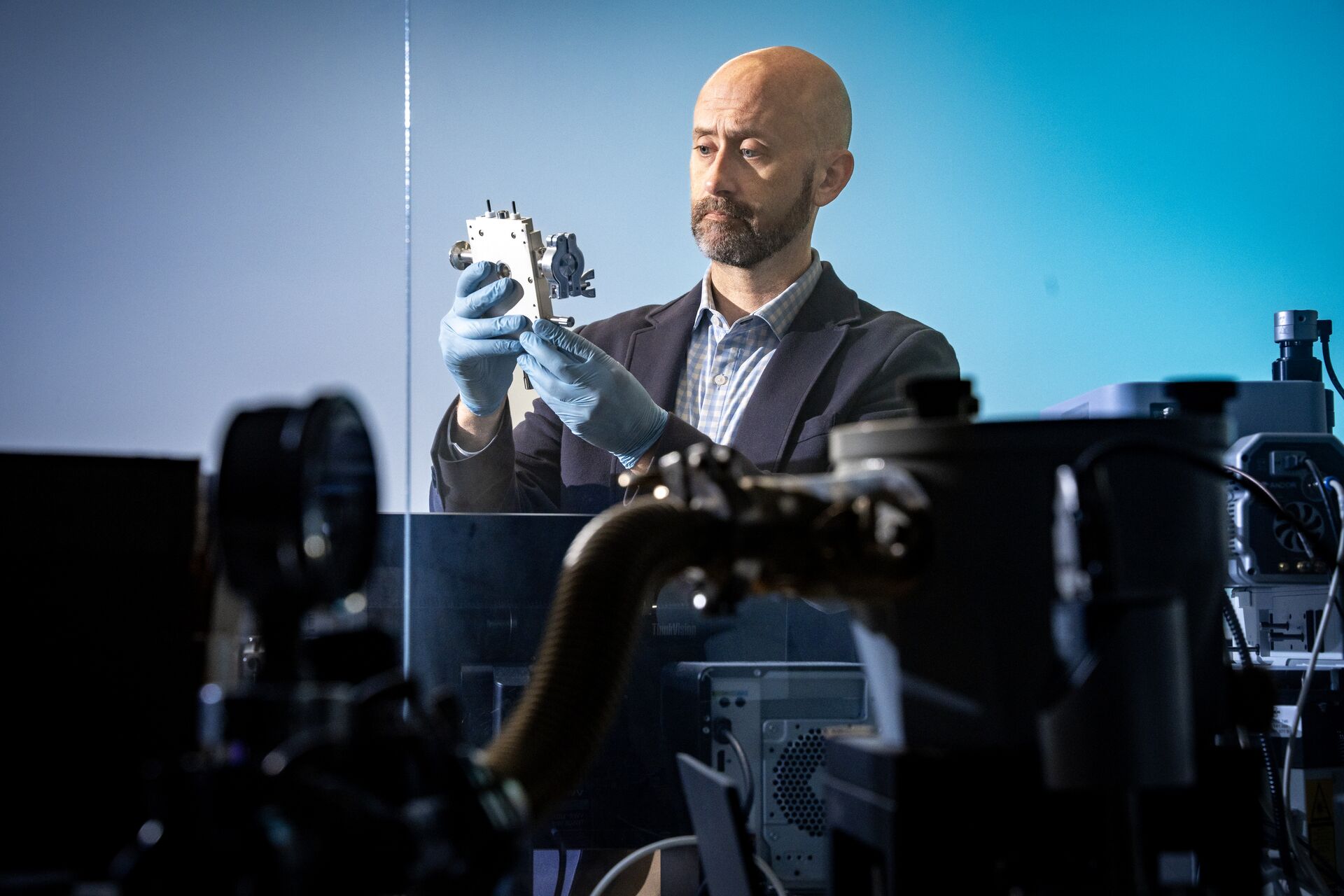 Person wearing blue gloves examines a small mechanical component in a high‑tech lab with equipment in the background.