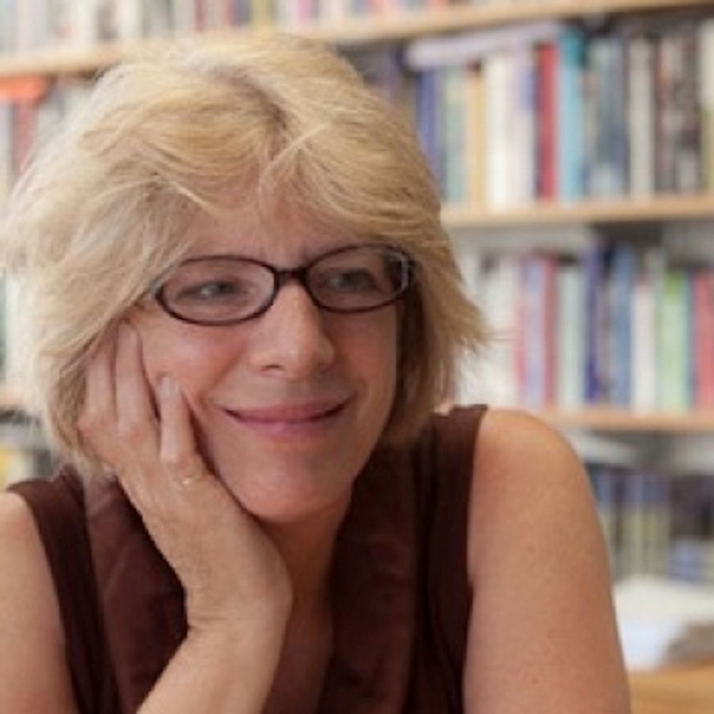 Person with light-colored hair and glasses, smiling softly with their head resting on one hand, seated in front of a bookshelf filled with books.