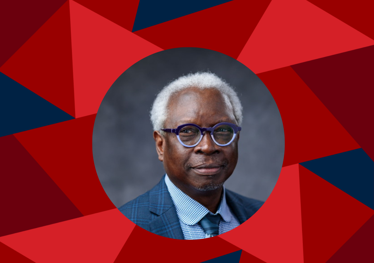 person with short white hair, glasses, and a blue plaid suit jacket, wearing a patterned tie and light blue checkered shirt, posed against a neutral gray background