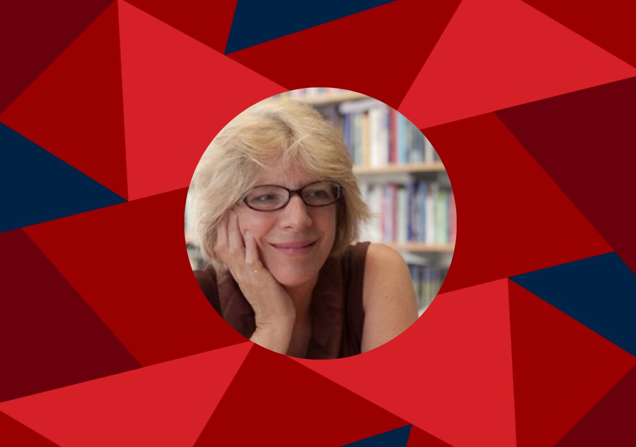 Person with light-colored hair and glasses, smiling softly with their head resting on one hand, seated in front of a bookshelf filled with books.