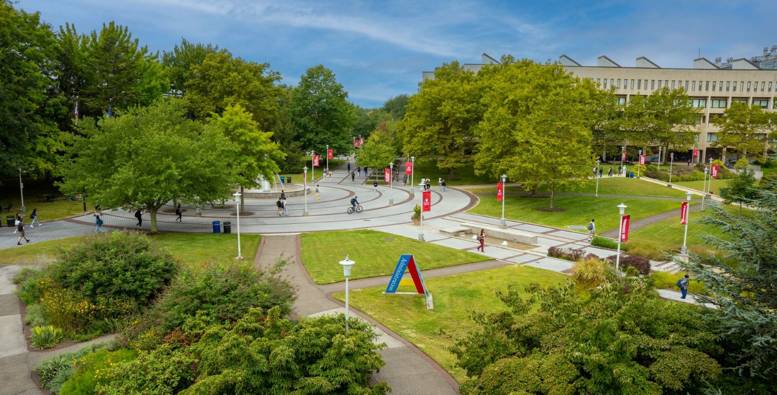 Here’s clear, accessible alOpen university courtyard with circular walkways, trees, and people walking and biking, with red Stony Brook University banners and a large academic building in the background.