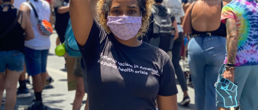 A masked protester raises a fist at a demonstration, wearing a shirt that reads “Systemic racism in America is a public health crisis.”