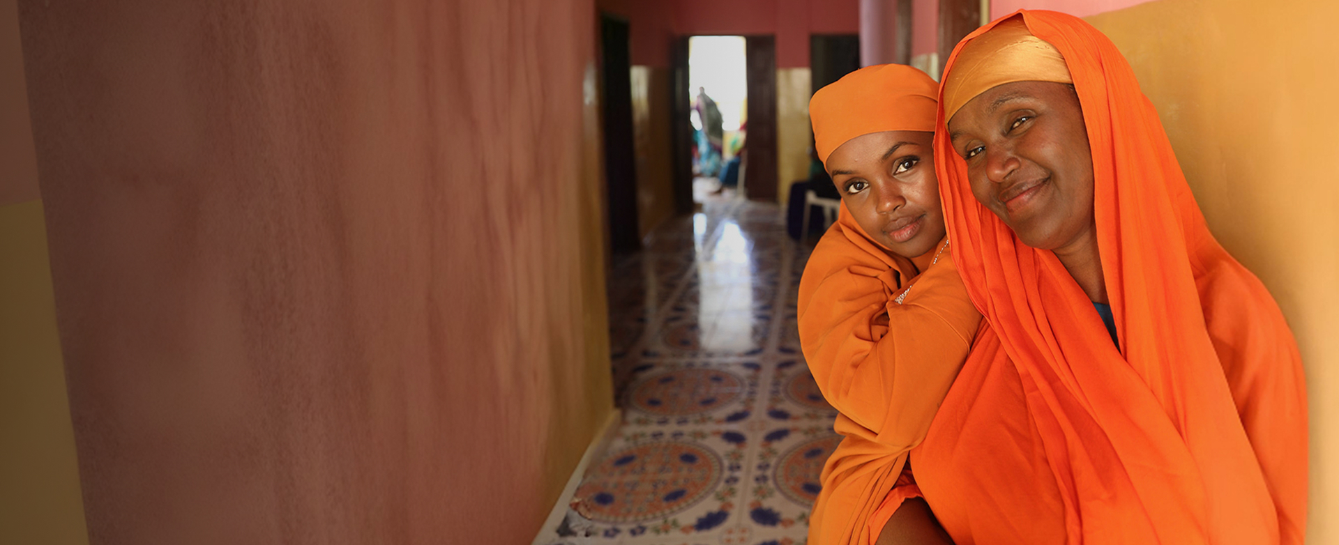 A young woman and an older woman in orange headscarves embrace in a tiled hallway, smiling at the camera