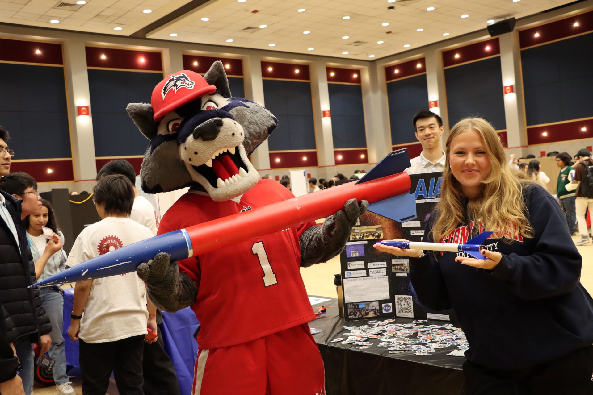 SBU mascot Wolfie and a student holding model rockets in a large multipurpose room.