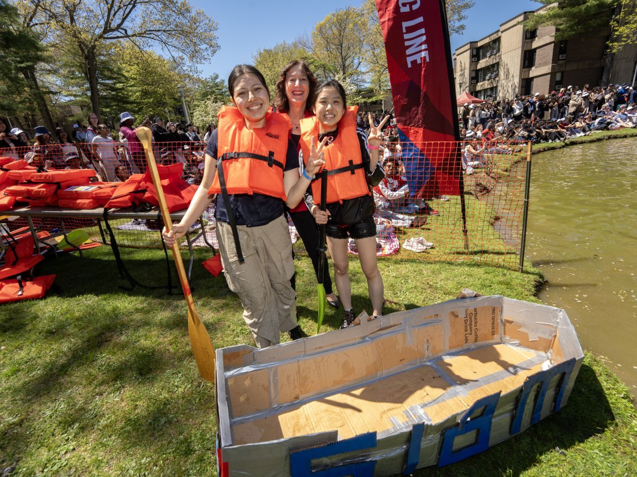 Two students smiling and wearing life vests next to a small, cardboard boat.